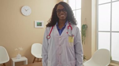 A smiling young african american woman doctor with curly hair and glasses, wearing a white coat and stethoscope, standing indoors in a hospital waiting room giving a thumbs-up gesture.