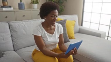 Orta yaşlı Afro-Amerikalı bir kadın, sarı yastıklı bir koltukta oturuyor, modern oturma odasında mavi bir tabletle etkileşime geçiyor, beyaz bir arka planı var.
