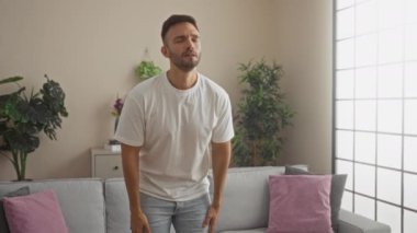 Genç İspanyol bir adam oturma odasında beyaz bir tişört ve kot pantolon giyerek oturur ve arka planda bitki ve yastıklarla çeşitli duygular gösterir..