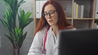 Kızıl saçlı ve gözlüklü odaklanmış bir kadın doktor klinikteki bir bilgisayarda çalışıyor, profesyonelliği ve sağlık hizmetlerini çağrıştırıyor..