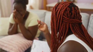 Bir kadın oturma odasında başka bir kadınla konuşuyor, elinde bir kağıt ve kalem tutarken sevgi ve aile bağını gösteriyor, kapalı alanda bir tartışma anı yakalıyor.