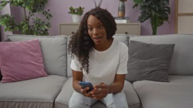 Genç, güzel, kıvırcık saçlı Afro-Amerikan bir kadın oturma odasında bir koltukta oturur, gülümser ve akıllı telefonuna bakar. Etrafı bitkiler ve dekorlarla çevrilidir..