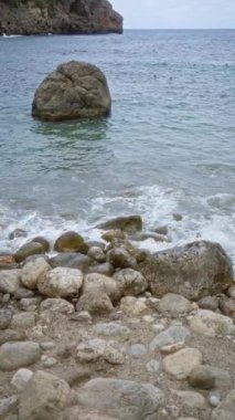 Kayalık plajlı kıyı şeridi ve Mallorca 'daki Cala Deia' nın turkuaz sularında büyük kaya tabakası bulutlu bir gökyüzünün altında İspanya.