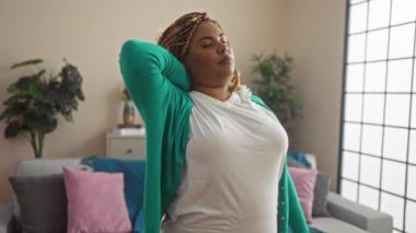 Saçları örgülü ve yeşil hırkalı güzel bir Afrikalı Amerikalı kadın oturma odasında boynunu tutarak boyun ağrısına işaret ediyor..