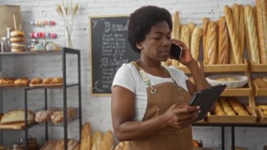 Fırında duran kıvırcık saçlı bir kadın elinde tabletle telefonda konuşuyor. Etrafı ekmek ve hamur işleriyle çevrili..