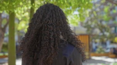 Güzel, kıvırcık saçlı Afro-Amerikan bir kadın şehir parkının dışında duruyor, şehir binalarının zeminine kurulmuş, yemyeşil, sıcak ve kendine güvenen.