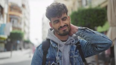 Sakallı genç bir adam rahatsızlık içinde, kot ceket ve sırt çantası giyiyor..