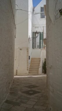 Locorotondo, İtalya 'da güneşli bir günde yeşil kepenkli kapıya çıkan merdivenleri ve beyaz taş binaları olan dar, kaldırımlı bir sokak..