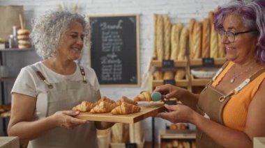 Fırında çalışan olgun kadın fırıncılar, biri ekmek ve hamur işleriyle çevrili bir tepsi kruvasanı diğerine uzatıyor..