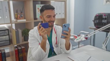 Genç İspanyol erkek veteriner, bir veteriner kliniğinde gülümserken akıllı telefon ve kredi kartı kullanıyor..
