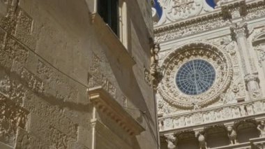 Derste, puglia, İtalya 'daki tarihi bir barok kilisesinin karmaşık cephesinde ayrıntılı taş oymalar ve açık mavi gökyüzünün altında geniş dairesel bir vitray bulunuyor..
