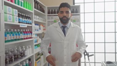 Gülümseyen genç İspanyol erkek eczacı modern eczanede ilaç raflarıyla duruyor..