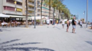 Güneşli bir açık hava geçidi boyunca palmiye ağaçları, binalar ve kafelerle kaplı yürüyen insanların bulanık görüntüsü, rahat bir yaz ortamı yaratıyor..