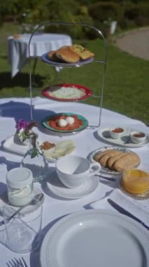 Ekmek, salata, peynir, badem, yoğurt, meyve suyu ve çay ile beyaz masa örtüleri ve çiçeklerle süslenmiş bir masaya kurulmuş açık hava kahvaltısı..