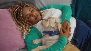 Saçları örgülü genç Afro-Amerikalı kadın rahat bir oturma odasında evcil köpeğiyle kanepede uyuyor..