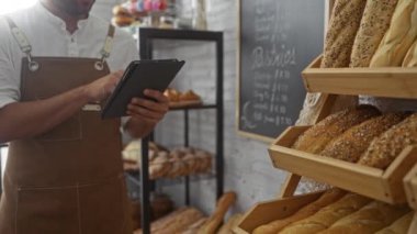 Fırında kahverengi önlük giyen genç adam tablet kullanıyor. Etrafı taze ekmek ve fırın ürünleriyle çevrili.