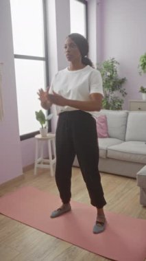 Genç bir Afrikalı Amerikalı kadın evinin oturma odasındaki pembe yoga minderinde çömeliyor..