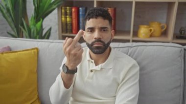 Impolite expression at home, young hispanic man boldly showing off rude middle finger, saying 'fuck you' with an unexpected show of defiance
