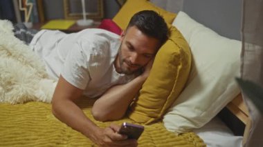 Yakışıklı, sakallı, genç bir İspanyol evdeki rahat bir yatak odasında yatıp cep telefonuyla meşgul oluyor..
