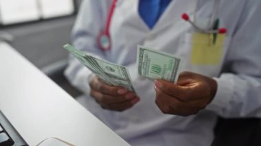 Klinik ortamında, laboratuvar önlüğü giyen Afro-Amerikalı bir kadın sağlık ortamında finansal işlemleri gösteren bir yığın Amerikan doları sayıyor..