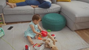 Tabletten kitap okuyan bir kadın, bebek oturma odasında oyuncaklarla oynarken, evde geçirdiği aile zamanlarını resmediyor..