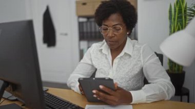 Ofis ortamında tablet kullanan bir kadın, yetişkin hayatının yarısında kıvırcık saçlı, gözlüklü ve beyaz gömlekli bir masada bilgisayarla çalışıyor..