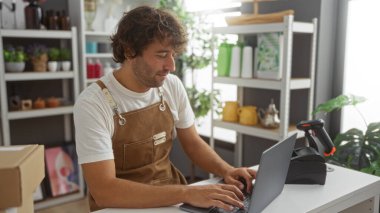 Dizüstü bilgisayarda çalışan genç bir adam dekoratif eşya raflarıyla iç dekorasyon mağazasında çalışıyor..