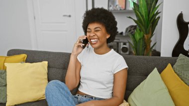 Dekoratif yastıklar ve gitar eşliğinde rahat bir oturma odasında telefonla konuşurken gülen Afrikalı Amerikalı kadın..