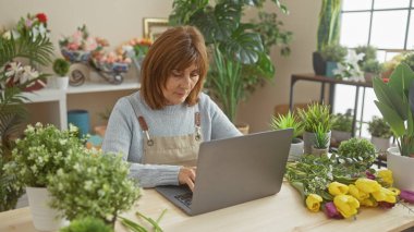 Çiçekçi dükkanında dizüstü bilgisayar kullanan olgun bir kadın. Etrafı bitkiler ve buketlerle çevrili. Girişimcilik ve çiçekçilikle dolu..