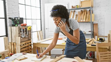 Afrikalı Amerikalı kadın bir atölyede birden fazla iş yapıyor, telefonda konuşuyor ve notlar alıyor.