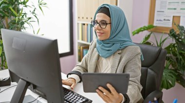 Modern ofis ortamında bilgisayar ve tabletle çalışan tesettürlü profesyonel bir kadın..