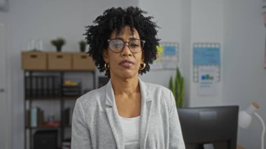 Kıvırcık saçlı ve gözlüklü güzel bir Afro-Amerikalı kadın arka planda rafları ve çizelgeleri olan kapalı bir ofiste güvenle duruyor..