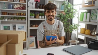 Sakallı genç adam elinde bir tablet tutarken gülümsüyor. Etrafı bitkilerle ve raflarda çeşitli dekoratif malzemelerle çevrili. Bu da bize, dost canlısı bir dükkan atmosferi olduğunu gösteriyor.