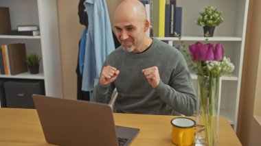Kel ve sakallı bir adam evde, dizüstü bilgisayarın önünde başarısını kutluyor. Samimi bir odada neşe ve zafer ifade ediyor..