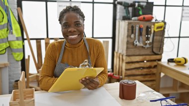 African american woman with braids in a carpentry workshop holding clipboard, smiling. clipart