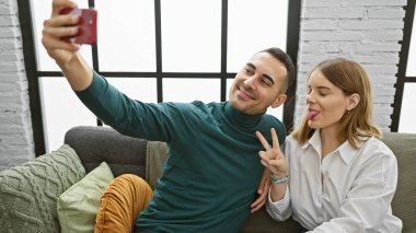 Bir kadın ve bir erkek, modern bir oturma odasında selfie için neşeli bir poz vererek, samimiyet ve mutluluğu paylaşıyorlar..
