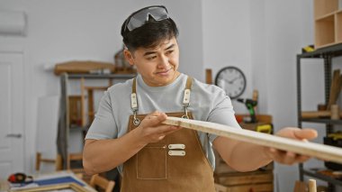 Genç Asyalı bir adam, iyi donanımlı bir marangozluk atölyesinde ahşap bir kalası inceliyor..
