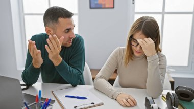 Stresli bir kadın ve hayal kırıklığına uğramış bir adam, modern ofis ortamında, masada dizüstü bilgisayar ve kırtasiyeyle sorunları tartışıyorlar..