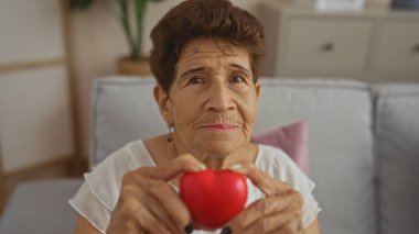Yaşlı İspanyol kadın elinde kırmızı kalp şeklinde bir nesne, rahat bir oturma odasında oturuyor..