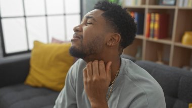 Düşünceli bir siyah adam evinin rahat iç kısmında oturmuş, günlük yaşamının samimi bir anını resmederken boynunu kaşır..