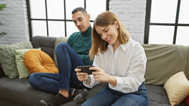 Bir kadın akıllı telefonunu koltukta oturan bir adamın yanında kontrol eder, modern yaşam tarzını ve kapalı mekandaki ilişkiyi tasvir eder..