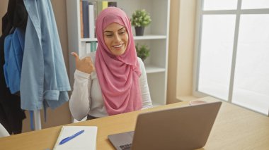 Başörtüsü takan gülümseyen bir kadın ev ofisinin dizüstü bilgisayarı ve masasında not defteri olan, pozitifliği ve verimliliği somutlaştıran bir ortamda başparmak kaldırıyor..