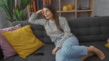 Düşünceli, genç bir İspanyol kadın, gri bir kanepeye uzanır. Etrafı renkli yastıklarla ve rahat bir iç mekânla çevrilidir..