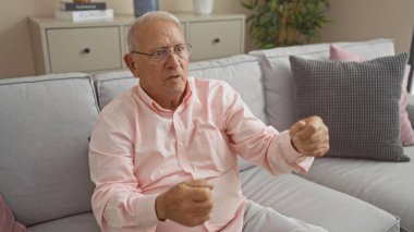 Yaşlı, beyaz gözlüklü, pembe gömlekli bir adam oturma odasındaki koltukta oturmuş sanki araba sürüyormuş gibi, yastıklı ve dolaplı ev dekorasyonu yapıyor..