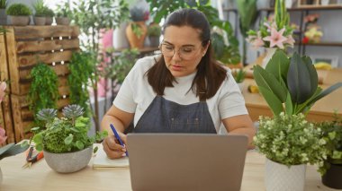 Çiçekçide yazan İspanyol kadın bitkiler, dizüstü bilgisayarlar ve gözlüklerle.