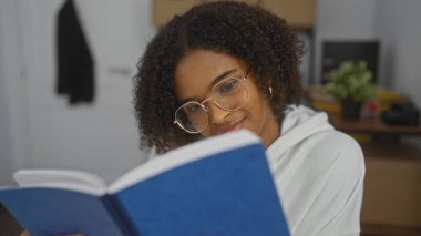 Güzel bir Afro-Amerikalı kadın kıvırcık saçlı, kapalı bir ofiste mavi kitap okuyor..