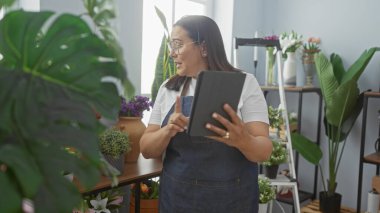 Çiçekçide tablet kullanan önlüklü İspanyol kadın. Etrafı çeşitli bitkiler ve iç dekorlarla çevrili..