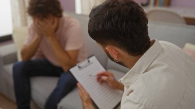 Oturma odasında terapi seansına katılan iki adam bir adamın not defterine not aldığı diğer adamın ise yüzünü kapadığı için üzgün olduğu görülüyor.