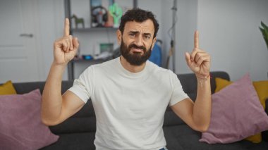 Beyaz tişörtlü orta yaşlı sakallı adam modern bir oturma odasında işaret parmaklarıyla şaşkın şaşkın bakıyor.