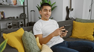 Modern bir oturma odasındaki gri bir kanepede uzanırken elinde tablet tutarak gülümseyen sıradan İspanyol bir adam..